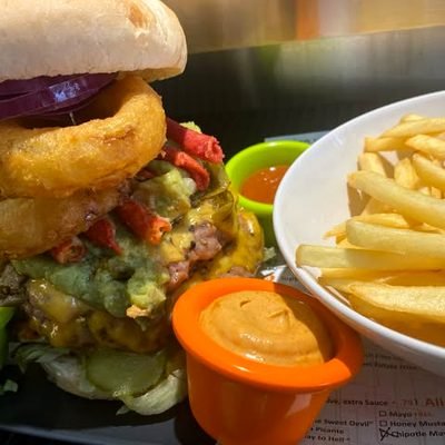 Burger mit Zwiebelringen, dazu eine Schale Pommes und zwei Dips.